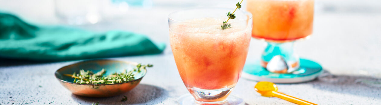 Glasses filled with a nectarine-hued Slushy Shirley cocktail garnished with a sprig of thyme sitting on a gray countertop.